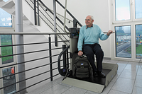 Älterer Herr sitzt entspannt auf einem Plattformlift im Treppenhaus eines modernen Gebäudes und lässt sich barrierefrei zwischen den Etagen transportieren.