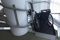 Plattformlift mit abgestelltem Rollstuhl auf einer Fahrplattform an einer kurvigen Innentreppe in einem modernen Gebäude.