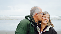 Older couple on the beach, the man lovingly kisses the woman on the cheek, symbolising joy of life and independence in later years